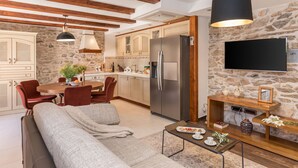 A modern kitchen with a large refrigerator, wooden cabinets, and a stone wall. There is a dining area with a table and chairs, and a living area with a sofa and a coffee table. - Josipovi Dvori in Skradin (Skradin)