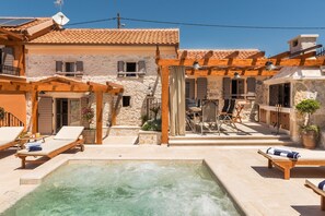 A pool area with lounge chairs, a pergola, and a stone building with a tiled roof.