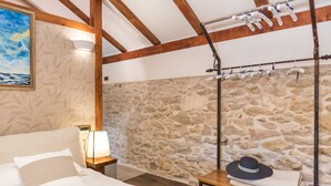 A bedroom with a stone wall, wooden beams, a bed with white linens, a nightstand with a lamp, and a hat on a small table. - Josipovi Dvori in Skradin (Skradin)