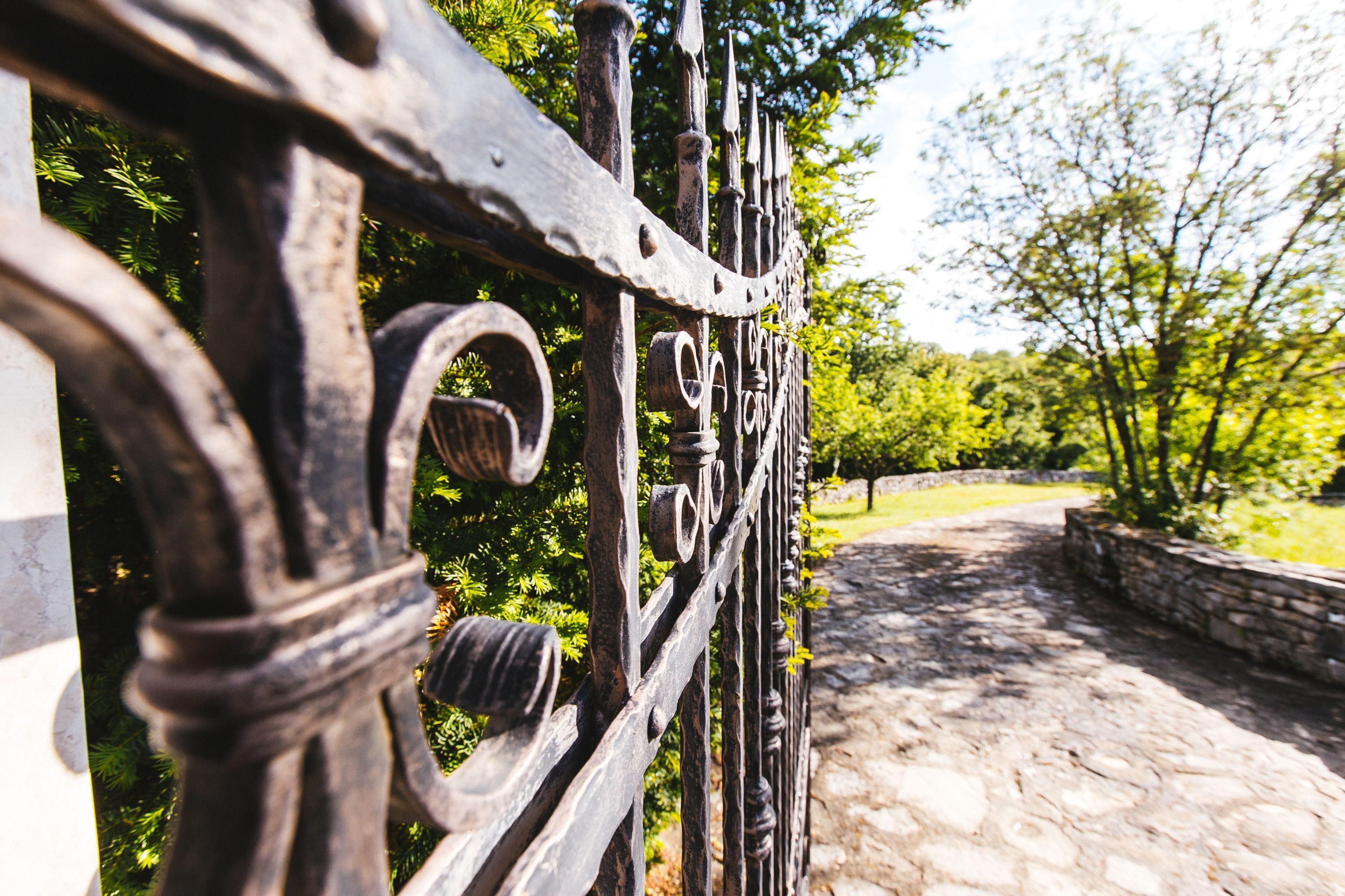 A wrought iron gate with intricate designs, leading to a stone pathway surrounded by greenery.