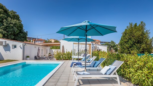 A swimming pool with lounge chairs and umbrellas, surrounded by greenery and buildings. - Amia in Pula With 3 Bedrooms and 2 Bathrooms (Pula)