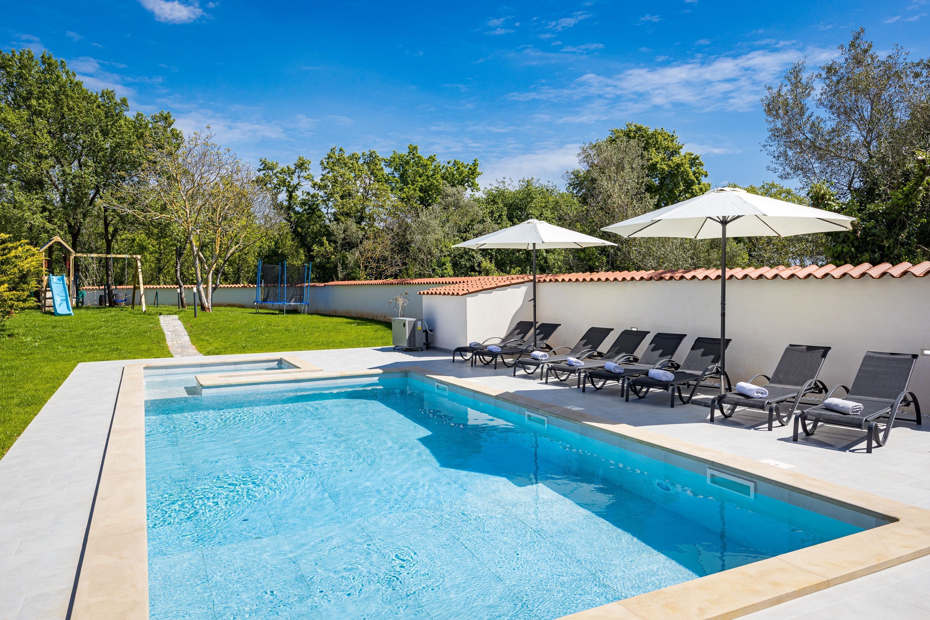 Una piscina con sillones reclinables y sombrillas, rodeada de un jardín y árboles.