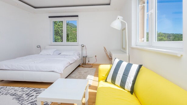 Un dormitorio con una cama, un sofá amarillo, una mesa blanca, una ventana con vistas a zonas verdes y una lámpara en el techo.