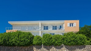 Un edificio moderno de dos plantas con balcones, un muro de piedra en primer plano y exuberantes arbustos verdes.