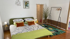 Een slaapkamer met een bed, een nachtkastje met een lamp, een wandklok, een lijst met een schilderij, een hangplant en een kledingrek.