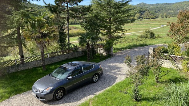Een auto geparkeerd op een grindpad, met een stenen muur en een hek aan de ene kant, en een uitzicht op een groen landschap met bomen en heuvels op de achtergrond.