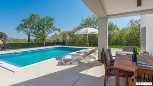 Piscina con sillas de descanso y una mesa, rodeada de un jardín y árboles.