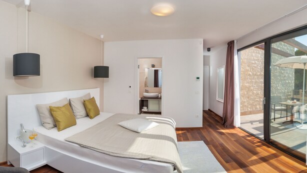 A modern bedroom with a large bed, bedside tables, a built-in sink, and a sliding glass door to a balcony. - Sunce in Okrug Gornji (Okrug)
