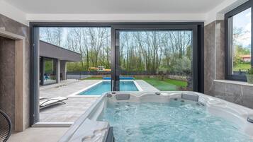 A modern indoor swimming pool area with a hot tub, large windows, and a view of an outdoor pool and greenery.