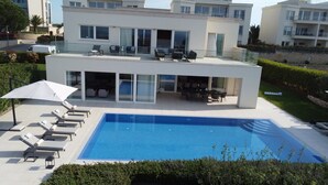 Une maison moderne à deux étages, dotée d’une piscine, d’un espace extérieur aménagé et d’un patio fermé par des baies vitrées.