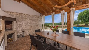 An outdoor patio with a stone fireplace, a wooden table, wicker chairs, and a view of a swimming pool. - Lanca in Sveti Lovre (Sveti Lovrec)