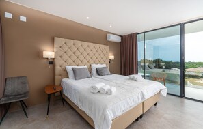 Quarto de hotel moderno com uma cama grande, uma mesinha de cabeceira, uma cadeira e uma janela ampla com vista para o exterior.