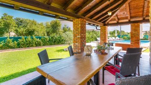 An outdoor patio with a wooden table and chairs, a brick pillar, and a view of a swimming pool. - Kamenica in Kras Krk (Dobrinj)