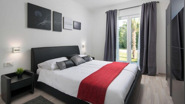 A modern bedroom with a large bed, a nightstand, a red throw blanket, and a view of the outdoors through a sliding glass door. - Deep Blue in Labin With 3 Bedrooms and 2 Bathrooms (Labin)