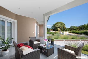 A covered patio area with wicker furniture, a table with a vase and a book, and a view of a landscaped garden and trees. - Deep Blue in Labin With 3 Bedrooms and 2 Bathrooms (Labin)