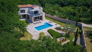 Une maison moderne à deux étages, dotée d’une piscine, entourée d’une végétation luxuriante et d’un mur en pierre.