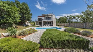Une maison moderne à deux étages, dotée d’une piscine, entourée de jardins bien entretenus et d’un mur en pierre.