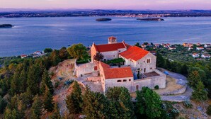 A historic stone building with a red roof, surrounded by trees and overlooking a large body of water with several islands. - Duzac in Kraj With 2 Bedrooms and 2 Bathrooms (Pasman)