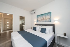 A modern bedroom with a large bed, a painting of sailboats, an air conditioner, and a bedside table with a lamp.