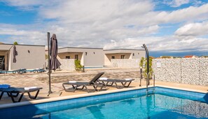 A swimming pool with lounge chairs and umbrellas, surrounded by buildings with stone walls and a clear sky. - M3 in Vir With 2 Bedrooms and 2 Bathrooms (Vir)