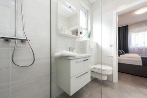 A modern bathroom with a shower, toilet, and sink, with a bed visible in the adjacent room.