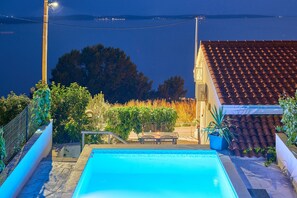 Piscina com vista para o mar, rodeada por vegetação e um edifício com telhado de azulejos.