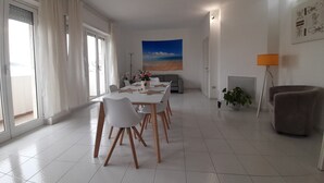 A spacious living room with a dining area, a sofa, and a large window with curtains. - Exclusive three-bedroom, three-bathroom apartment by the sea at Poetto (CAGLIARI)