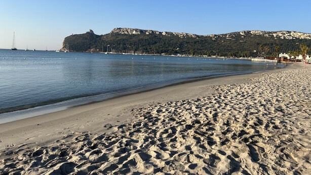 A sandy beach with a calm sea, a rocky hill in the background, and a few boats. - Exclusive three-bedroom, three-bathroom apartment by the sea at Poetto (CAGLIARI)