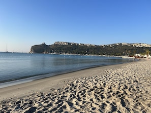 A sandy beach with a calm sea, a rocky hill in the background, and a few boats. - Exclusive 3-bedroom, 3-bathroom apartment on the beachfront in Poetto (CAGLIARI)