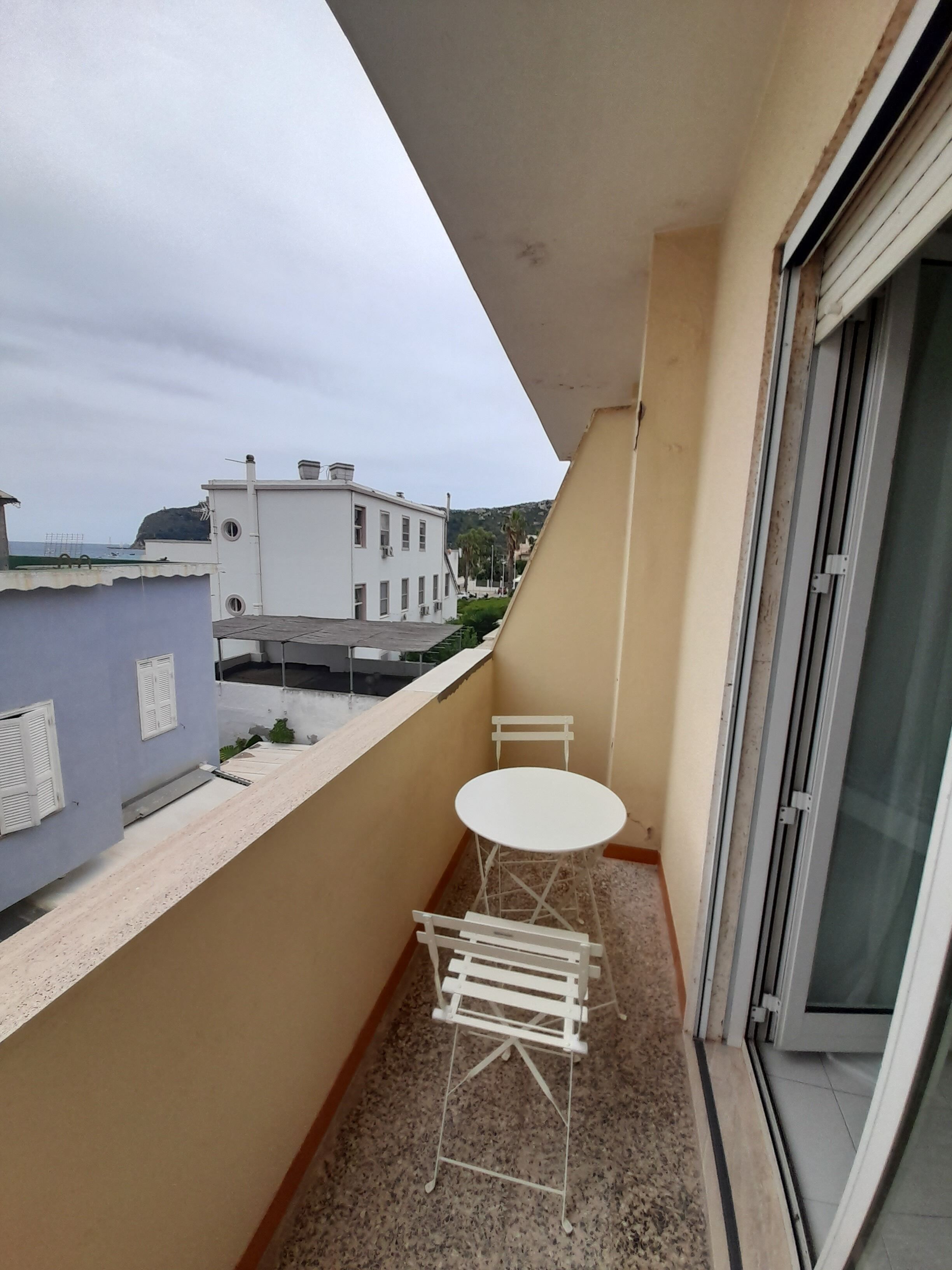 Un petit balcon avec une table et des chaises blanches, une fenêtre et une vue sur des immeubles et une colline au loin.