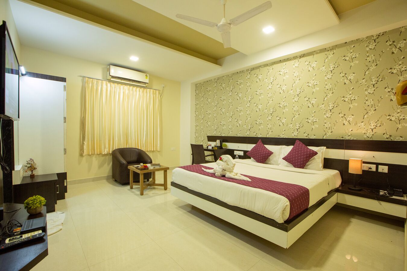 A hotel room with a bed, a desk with a computer, a chair, an air conditioning unit, and a ceiling fan.