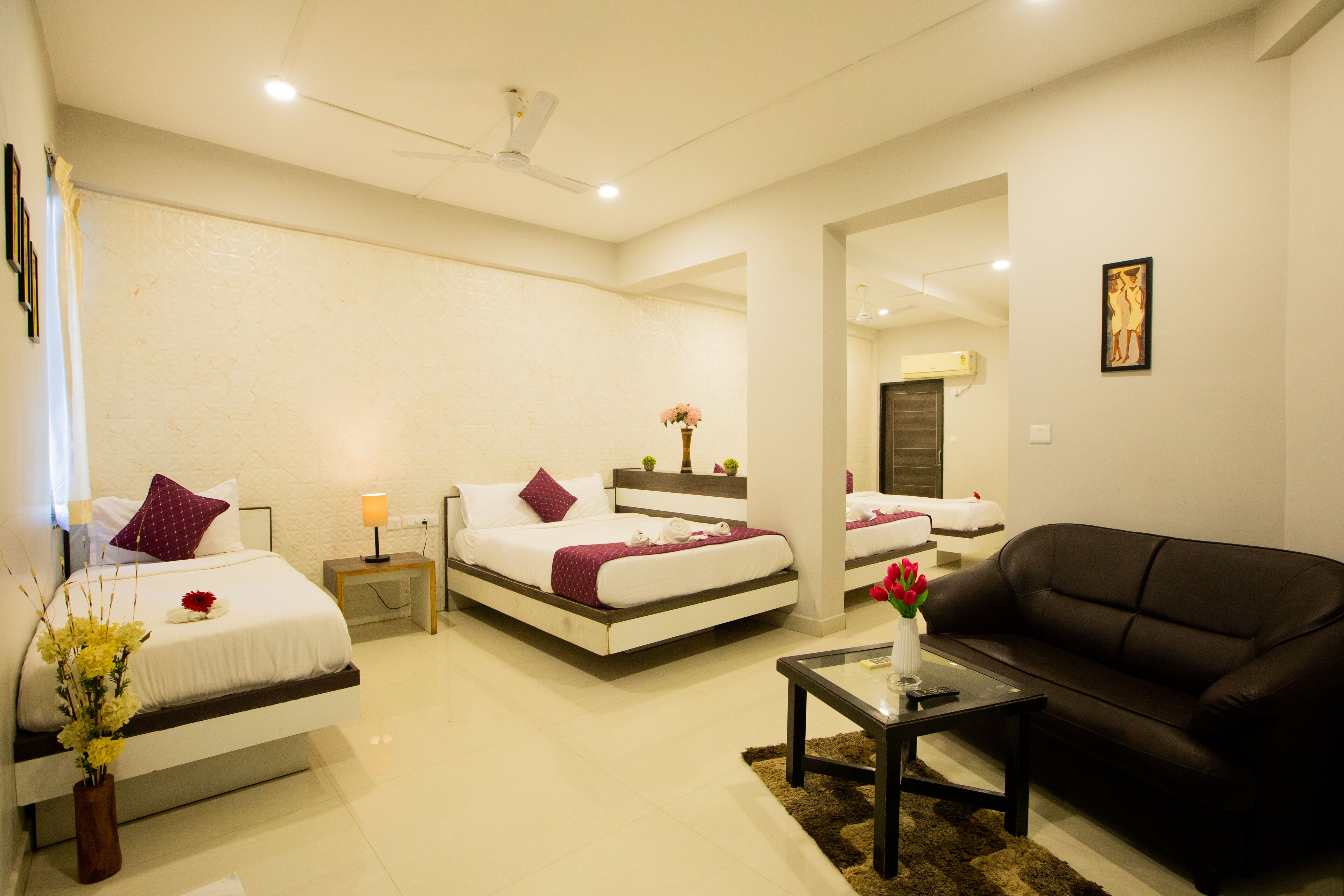 A modern hotel room with two beds, a black leather sofa, a glass coffee table, and a small side table with a lamp.