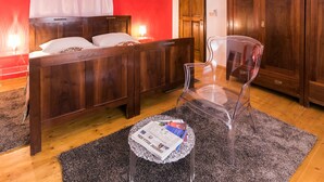 Une chambre avec un lit en bois, une chaise en plastique transparent, une petite table ronde sur laquelle repose un journal, et un mur rouge.