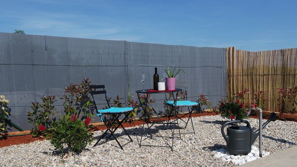 Un espace patio avec une table et deux chaises, une plante en pot, une bouteille de vin et un arrosoir.