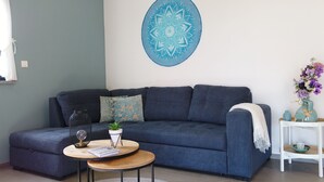 Un salon avec un canapé bleu foncé, une table basse ronde sur laquelle se trouvent un livre et un objet décoratif, une décoration murale et une table d’appoint avec un vase de fleurs.