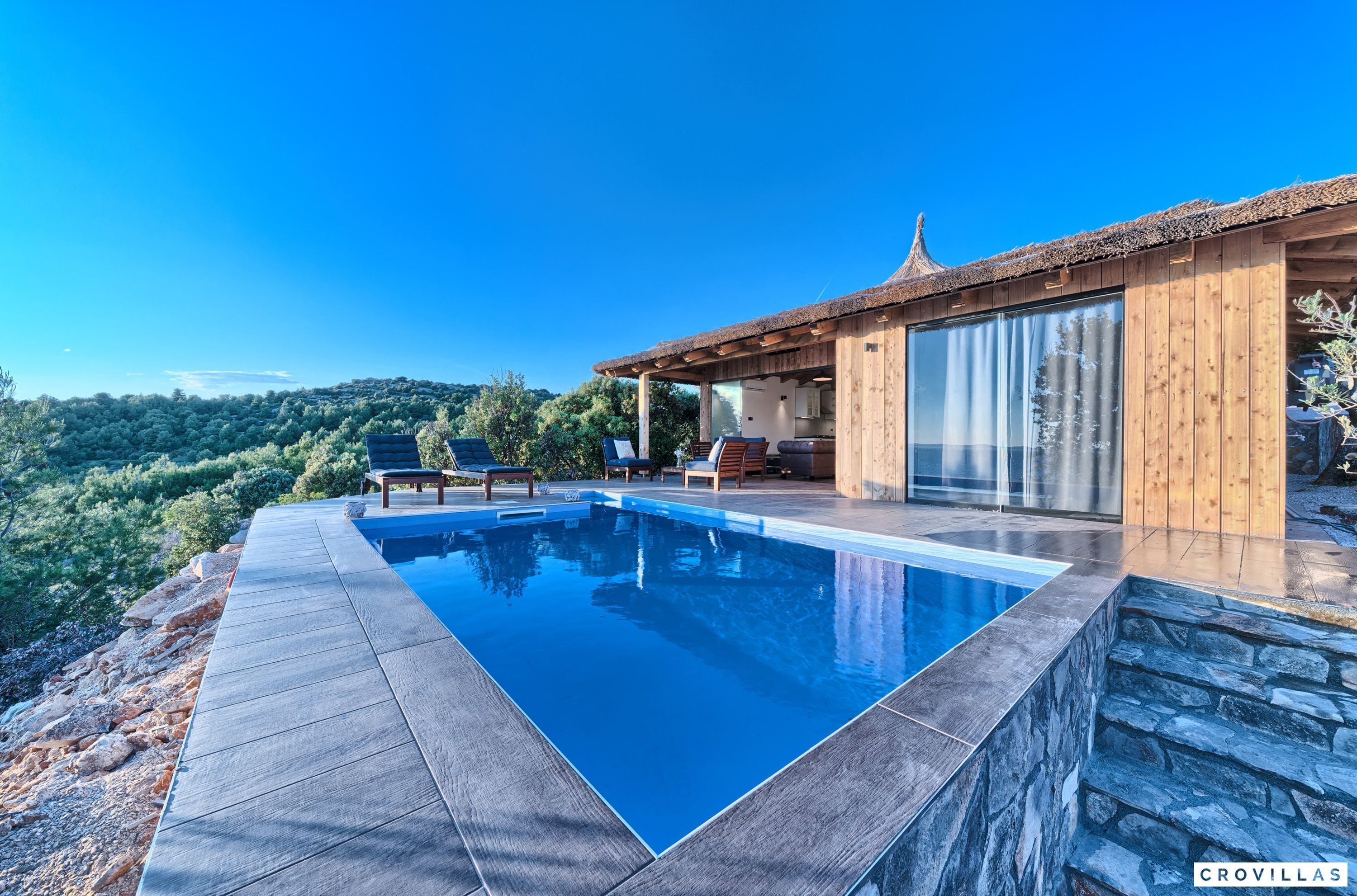 A modern villa with a swimming pool, wooden deck, and a view of the surrounding hills.