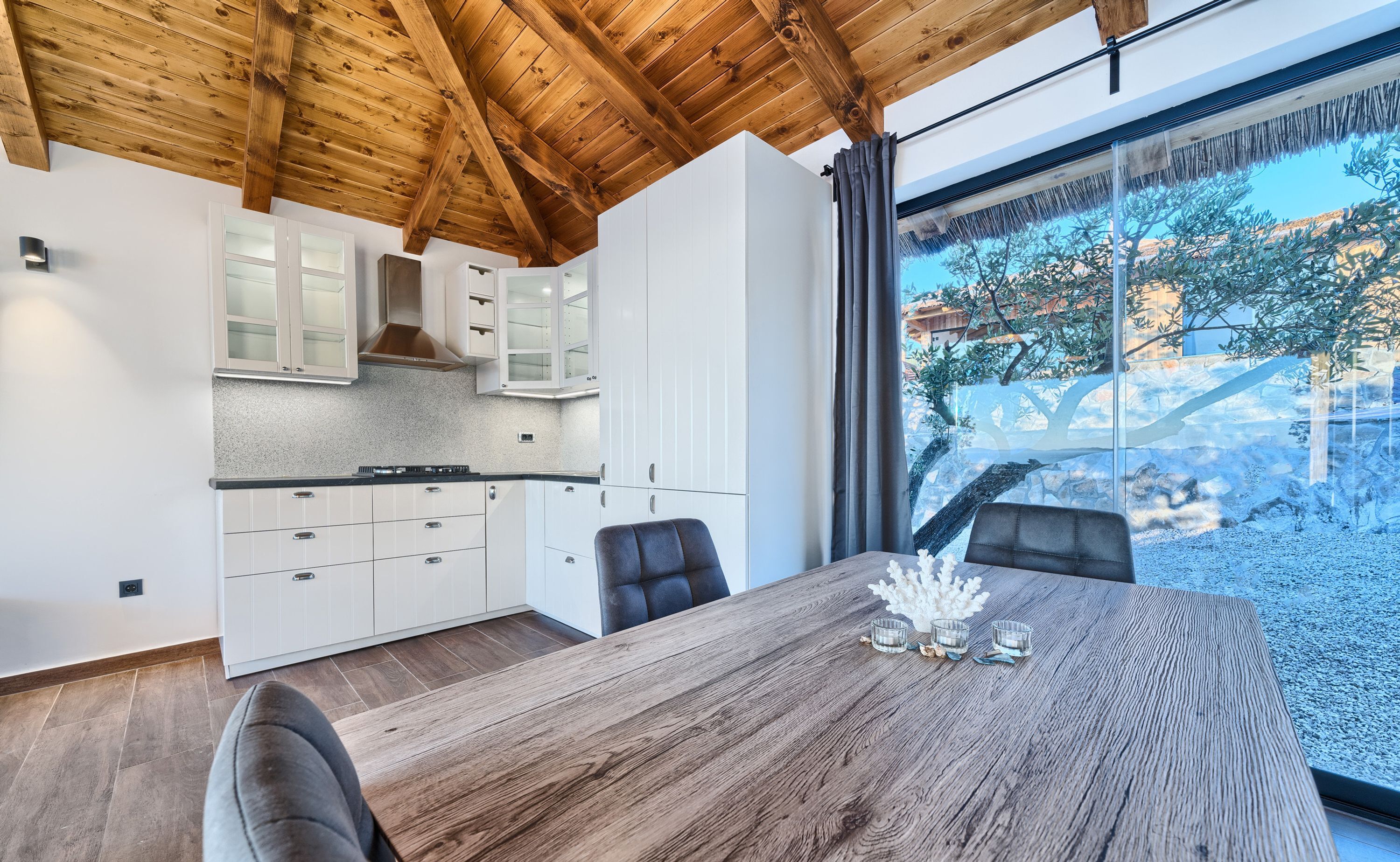 A modern kitchen with white cabinets and a wooden ceiling, a dining area with a wooden table and chairs, and a large window offering a view of trees and buildings.