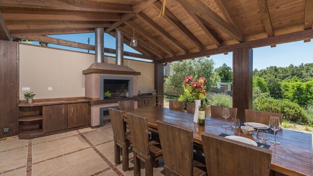 An outdoor kitchen area with a wooden table and chairs, a built-in oven, and a view of greenery. - Dali in Svetvin Enat (Svetvincenat)
