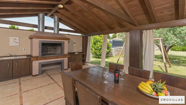 Ein Outdoor-Kochbereich mit Holzboden, einem eingebauten Ofen und einem Esstisch mit Obstschale und Weingläsern.