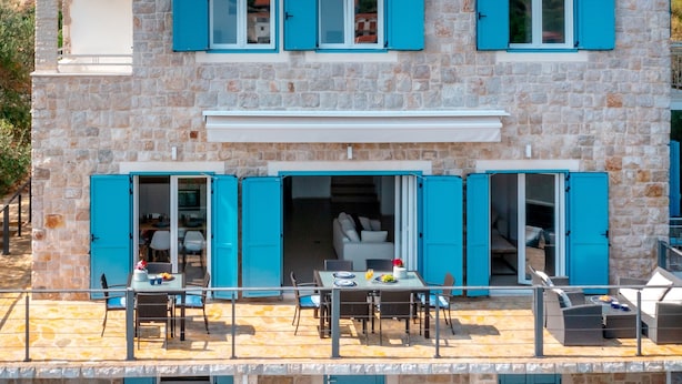 Un edificio de piedra con contraventanas azules, un balcón con mobiliario para exterior y un interior visible con comedor.