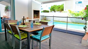 Een eetruimte met uitzicht op het zwembad, met een houten tafel, stoelen en een fles wijn.