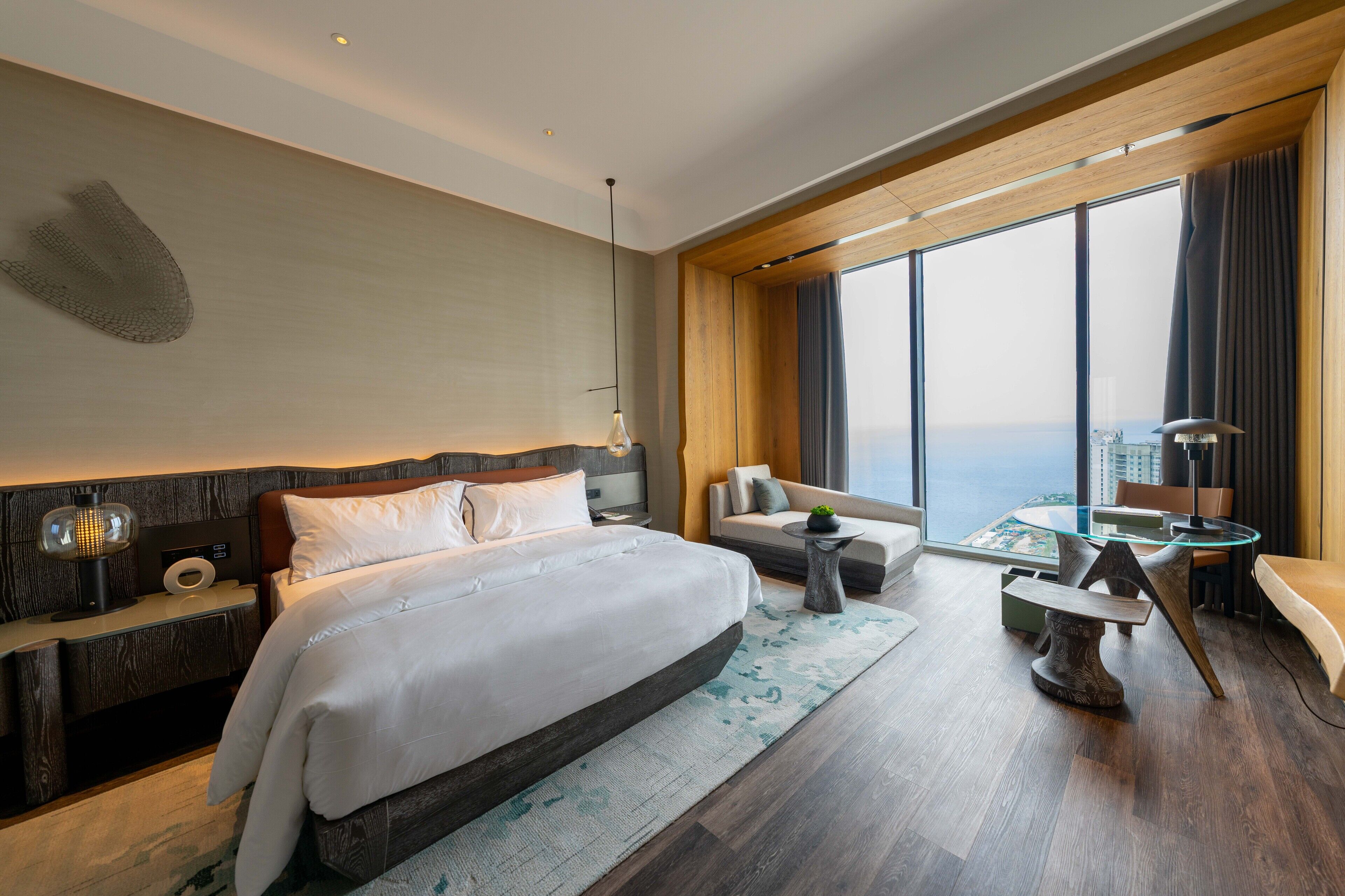 A modern hotel room with a large bed, a sofa, a glass table, and a view of the city.