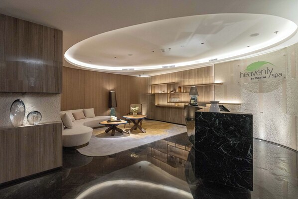 Interior moderno de spa con un mostrador de recepción, una zona de estar con sofá y una pequeña mesa, y una pared de madera con estanterías.