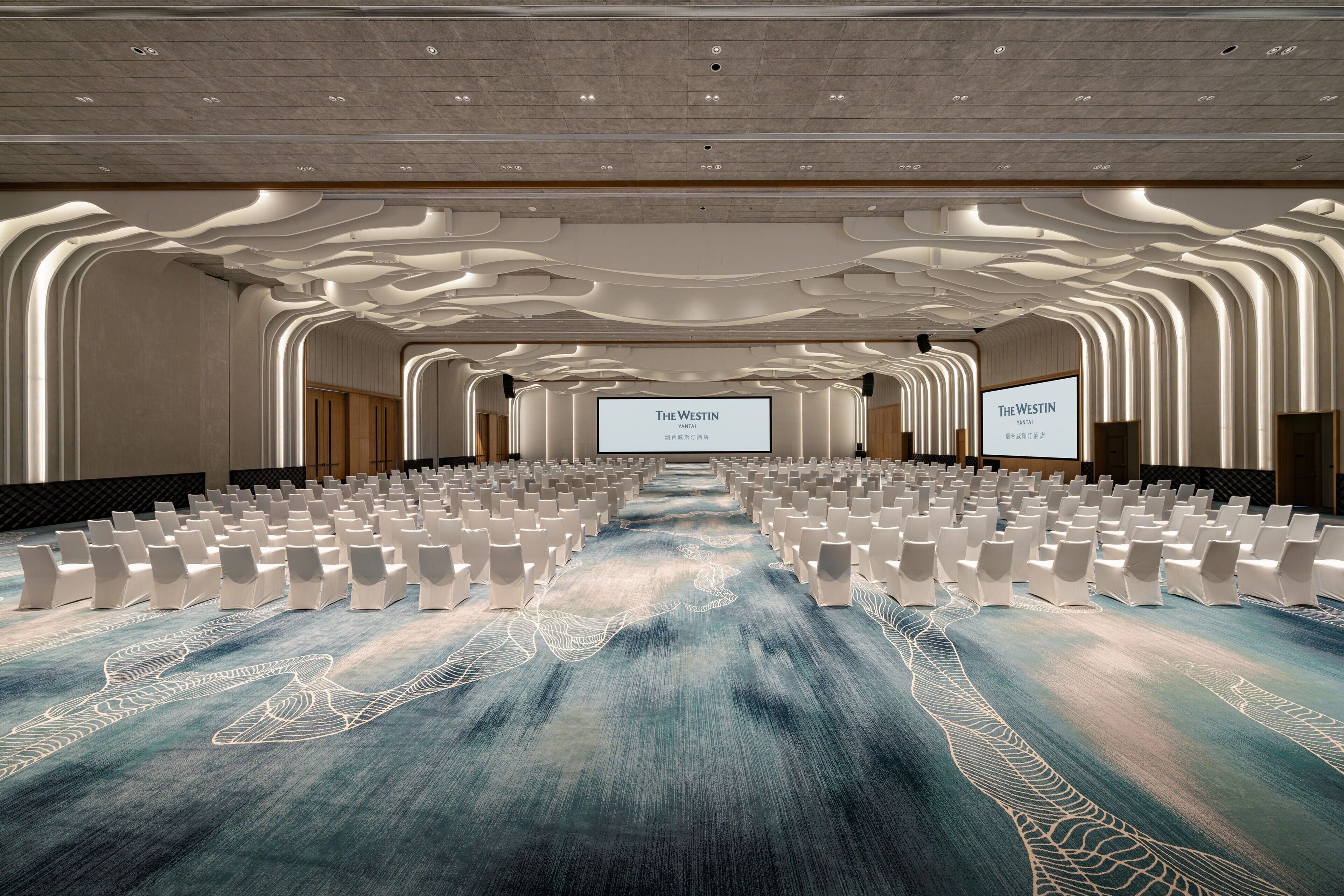 Un salón de conferencias con filas de sillas blancas, una pantalla grande mostrando 'The Westin' y una alfombra con un diseño ondulado.