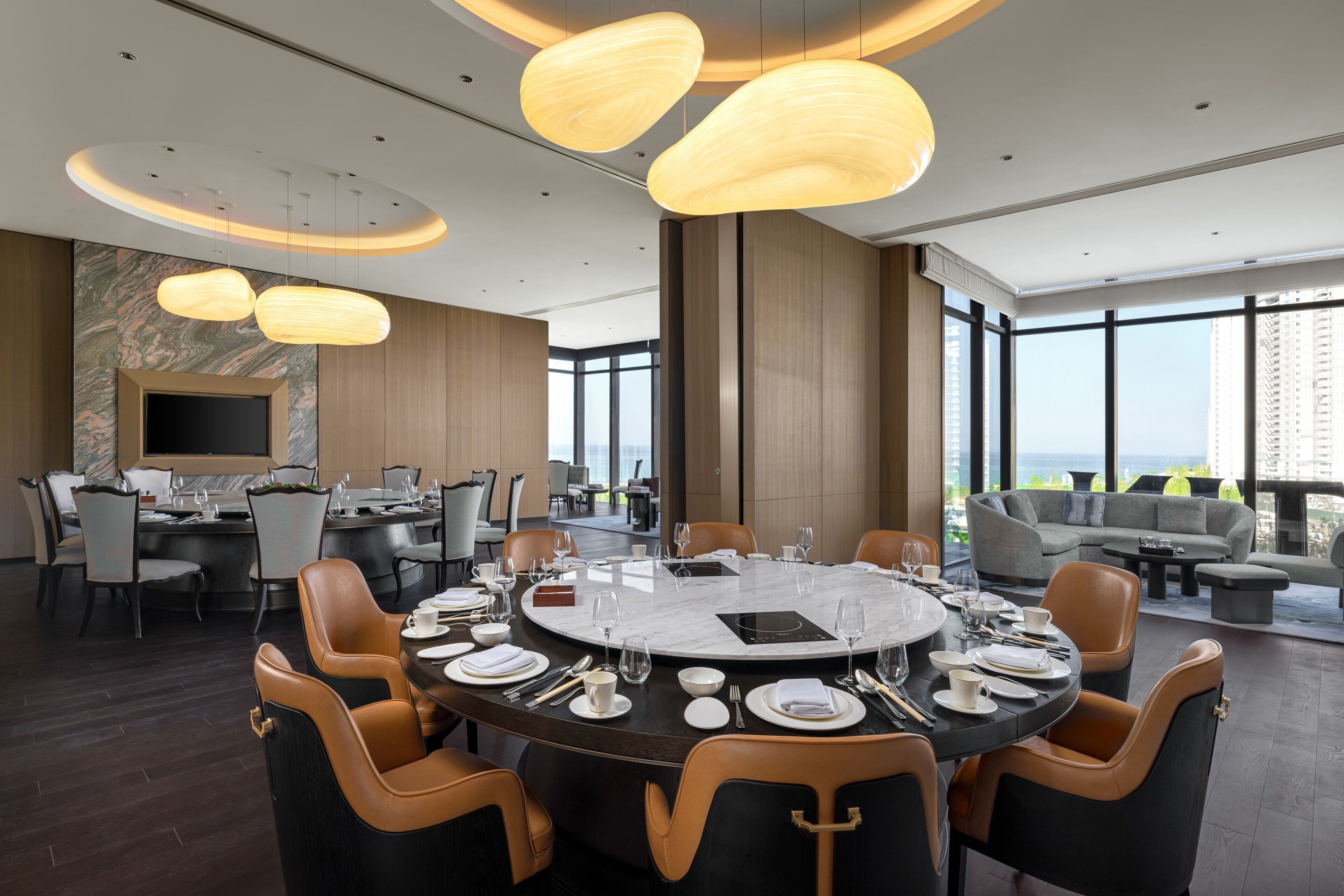 A modern dining area with a round table set for six, surrounded by leather chairs. There is a large window offering a view of the cityscape.