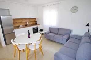 Een moderne keuken met witte kasten, een houten aanrecht en een eethoek met een witte tafel en stoelen. Er is een grijze bank en een klok aan de muur.
