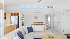 A spacious living area with a sofa, coffee table, and a dining table with chairs. There is a kitchen area with cabinets and a refrigerator. The room has a large window and a wall-mounted clock. - Unit 6 'Sea Breeze Villas' (Coolum beach)