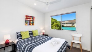 Quarto com cama, duas mesas de cabeceira, uma cadeira, ventilador de teto, janela com vista para uma casa e piscina, e uma pintura abstrata colorida na parede.