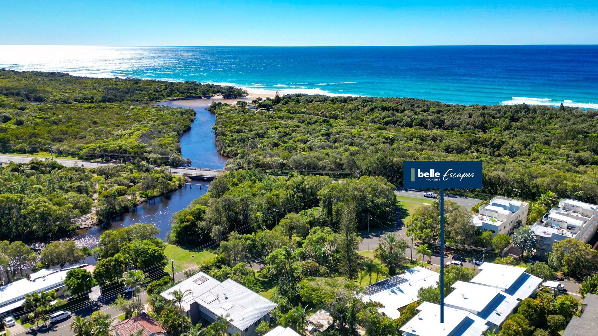 A residential area with a river, a bridge, and a beach in the background. There is a sign for 'Belle Escapes Property'.