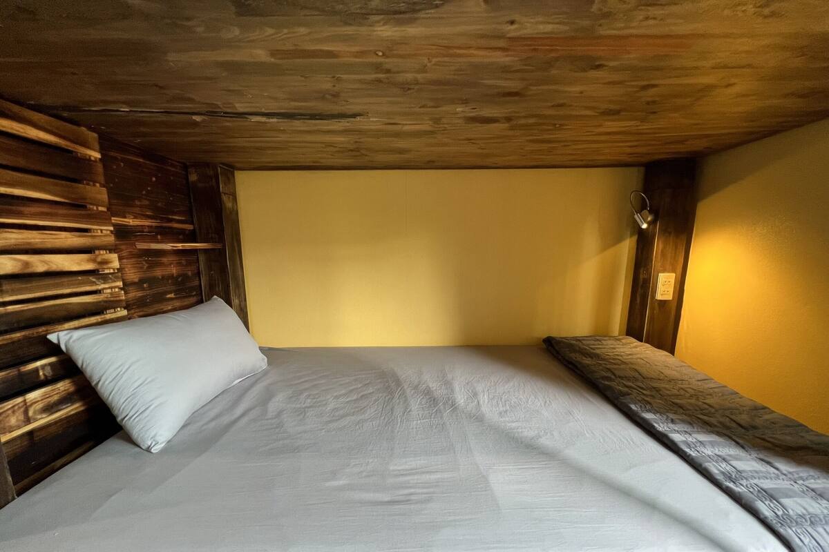 A single bed with a wooden headboard and a plain mattress in a room with yellow walls and a wooden ceiling.